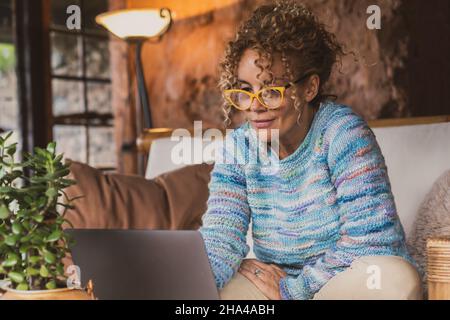 Porträt einer glücklichen modernen Frau zu Hause, die mit dem Computer auf dem Sofa sitzt und eine Brille trägt. Weibliche Menschen surfen im Internet und arbeiten lächelnd mit einem Laptop. Indoor-Technologie Freizeitangebot Konzept Stockfoto