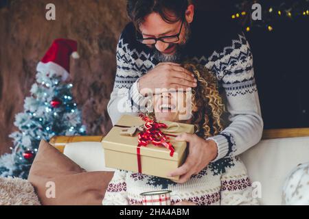 Mann überraschen eine Frau, die hier die Augen schließt und ein weihnachtsgeschenk für die Weihnachtsfeier zu Hause gibt. Menschen genießen den Urlaub zu Hause mit Spaß und Glück. Überglücklich Frau erhalten Geschenk Stockfoto