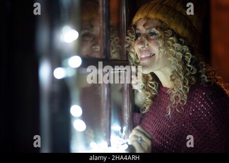 Glückliche junge Erwachsene Frau schauen vor den Fenstern zu Hause in der Nacht. Weibliche Menschen mit lockigen Haaren und Strickmütze genießen innen in der Wintersaison. Weihnachtsbeleuchtung und Reflexion auf dem Glas Stockfoto