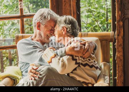 Ältere Männer und Frauen in Liebe und Zärtlichkeit zu Hause. Ältere ältere ältere Paare genießen die Beziehung umarmen und füreinander auf dem Sofa sitzen im Wohnzimmer mit Blick im Freien Stockfoto