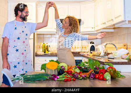 Fröhliche Erwachsene Paar tanzen und Spaß haben zusammen in der Küche zu Hause, während die Zubereitung von gesundem Gemüse auf dem Tisch. Überglücklich Frau und Mann in der Liebe bereiten Mittagessen und genießen Sie die Beziehung Stockfoto