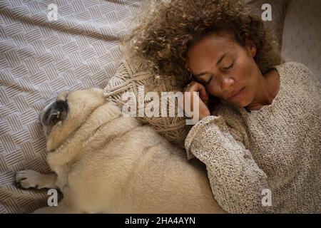 Junge Erwachsene Frau schlafen zu Hause im Bett mit ihrem Mops Hund zusammen auf dem Bett. Müde Frau mit Hund liegt auf dem Bett. Draufsicht auf müde Frau schlafen mit ihrem Hund auf gemütlichen Bett zu Hause Stockfoto