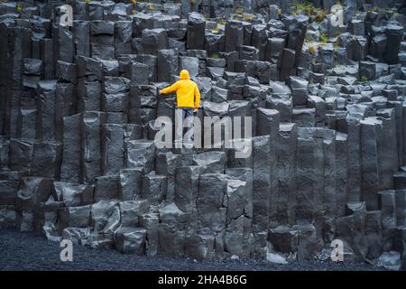 Mann mit gelbem Mantel und Blick auf reynisdrangar Basaltsäulen am Strand von Reynisfjara, island. Stockfoto