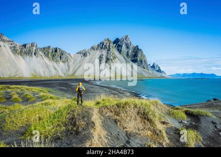 Mann mit Rucksack auf schwarzer Sanddüne auf der Stokksnes-Landzunge an der südöstlichen isländischen Küste mit vestrahorn-Sommerpanorama von island, europa. Luftpanorama. Stockfoto