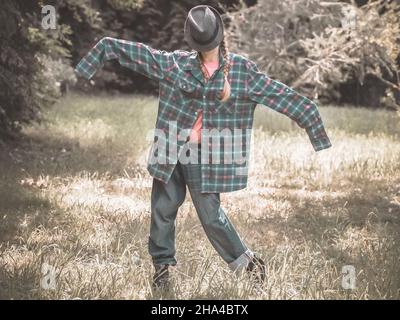 Eine Dame mit einem Hut im Gesicht und einem karierten Hemd mit übermäßig langen Ärmeln steht in einer Pose auf einem grünen Rasen. Parodie auf Gartenscheuche Stockfoto