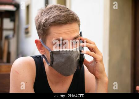 Auf der Pub-Terrasse befindet sich ein junger, hübscher weißer Mann mit braunen Haaren in schwarzem Unterhemd und grauer Gesichtsmaske Stockfoto