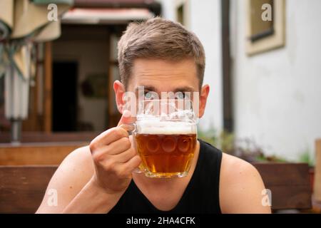 Junger, hübscher weißer Mann mit braunen Haaren in schwarzem Unterhemd und einem Pint Bier in der Hand auf der Pub-Terrasse Stockfoto