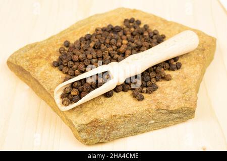 Viel ganz würziger schwarzer Pfeffer in einer Holzlöffel mit natürlichem Flachgestein auf einfachem Holz Stockfoto