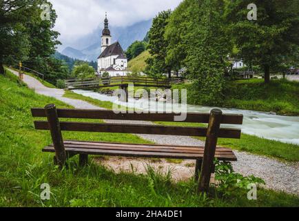 Eine Holzbank mit Pfarrkirche, Fluss abd Berge im Hintergrund. berchtesgaden, ramsau deutschland. Stockfoto