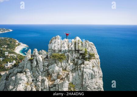 Luftaufnahme des Mannes Tourist in roter Jacke, die auf dem Felsen oben auf dem Katzenberg steht und seine Hände vor der Landschaft von simeiz cosatline hebt. krim. Stockfoto