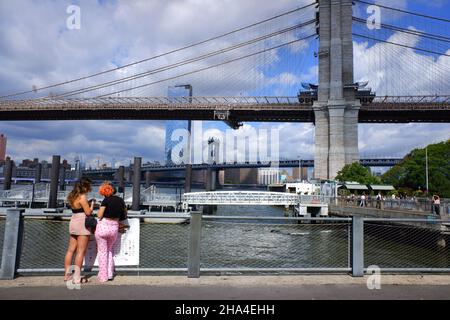 Brooklyn Bridge über East River mit Manhattan Bridge im Hintergrund zwei junge Frauen am Pier 1 im Brooklyn Bridge Park im Vordergrund.Brooklyn.New York City.USA Stockfoto