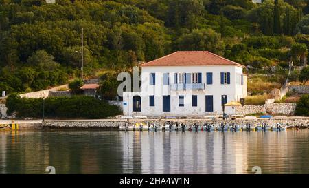 griechenland, griechische Inseln, ionische Inseln, kefalonia, fiskardo, Morgenstimmung, teilweise bewölktem Himmel, großes weißes Haus mit rotem Ziegeldach, davor befinden sich viele kleine Motorboote im Hafenbecken, das Haus spiegelt sich im ruhigen Wasser wider Stockfoto