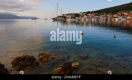 griechenland,griechische Inseln,ionische Inseln,kefalonia,fiskardo,Morgenstimmung,teilweise bewölktem Himmel,Blick von Osten über das Hafenbecken nach fiskardo. Im Vordergrund ein grün-blaues Meer mit Felsen,rote Bojen in der mittleren Distanz,ein großes Segelboot und andere Segelboote stehen vor Anker Stockfoto