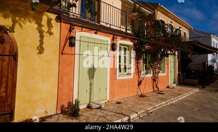 griechenland, griechische Inseln, ionische Inseln, kefalonia, fiskardo, Morgenstimmung, teilweise bewölktem Himmel, Häuserreihe, gelb, grün und ocker im Morgenlicht, Schatten einer Laterne auf einer grünen Tür, davor ein Bürgersteig, an der oberen rechten kleinen Ecke mit einem blauen Himmel Stockfoto