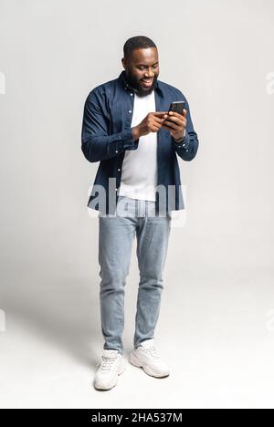 Fröhlicher und unbeschwerter afroamerikanischer Mann mit Smartphone isoliert auf grau, schwarzer Kerl verbringt Freizeit in sozialen Netzwerken, online chatten, einkaufen, mit neuen mobilen App, volle Länge, vertikal Stockfoto