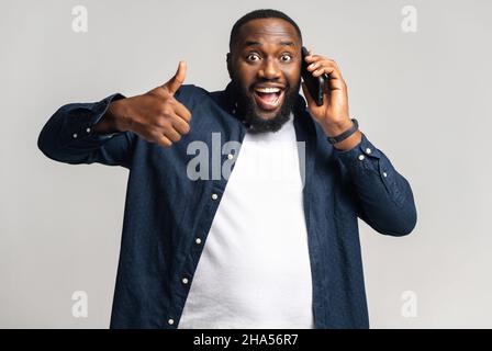 Porträt eines hübschen jungen afroamerikanischen Mannes, der Smartphone spricht und den Daumen nach oben zeigt, Kerl, der die Kamera anschaut und lächelt, fühlt sich zufrieden, isoliert Stockfoto