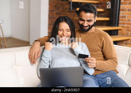 Fröhliches indisches Paar, das auf den Laptop-Bildschirm schaute und lächelte. Überglücklich freute Frau beim Online-Shopping mit ihrem Mann. Geld überweisen, Online-Banking-Konzept Stockfoto