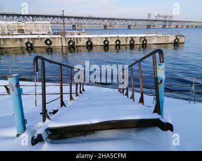 Winteransicht der verlassenen Flusspiers am Dnjepr. Rostige Poller im Schnee. Die Piers sind mit Schnee bedeckt. Stockfoto