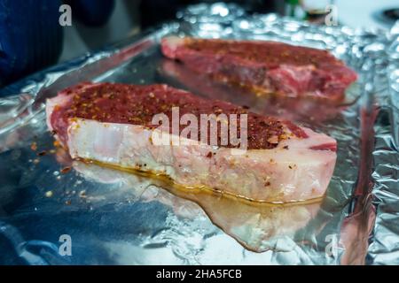 Abgewinkelte Ansicht von zwei großen, rohen Steaks, frisch gewürzt auf einem Backblech, das mit Zinnfolie bedeckt ist Stockfoto