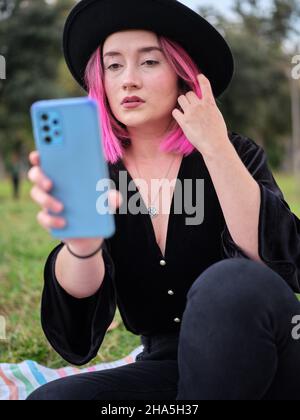 Frau posiert, während sie auf ihr Mobiltelefon schaut und ihr rosafarbenes Haar berührt Stockfoto