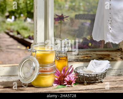 Zwei mit Echinacea-Salbe gefüllte Vorratsgläser und eine Glaskaraffe aus Pflanzenöl stehen zusammen mit einem Metallkorb und textilen Taschentüchern auf einem Holztisch vor einem Werkstattfenster Stockfoto