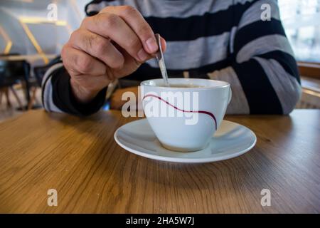 Junger Mann, der in einem modernen Café sitzt und Kaffee rührt. Stockfoto