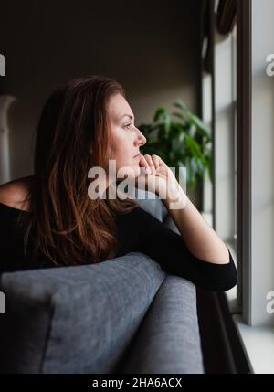 Frau schaute nachdenklich aus dem Fenster, während sie auf dem Sofa saß. Stockfoto