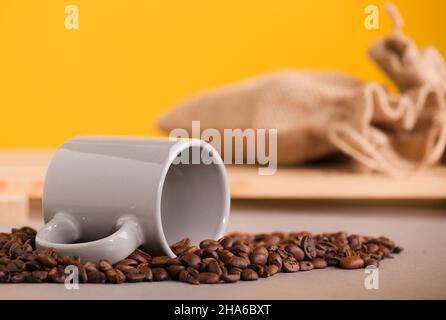 Graue Tasse Kaffee und geröstete Kaffeebohnen, Bast-Säcke mit gelbem Hintergrund Stockfoto