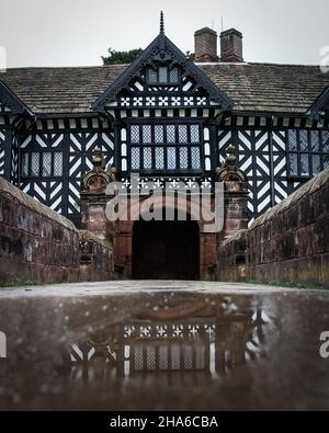 Die Spiegelung einer Pfütze, die die Tudor-Fassade einfängt. Speke Hall ist ein holzgerahmtes Tudor-Herrenhaus in Speke, Liverpool, England. Stockfoto