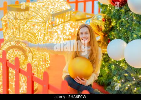 Lächelnde Dame mit einem riesigen Weihnachtsball, während sie neben einer festlichen Tanne und einem funkelnden dekorativen Zug sitzt. Festliche Neujahrsstimmung. Stockfoto