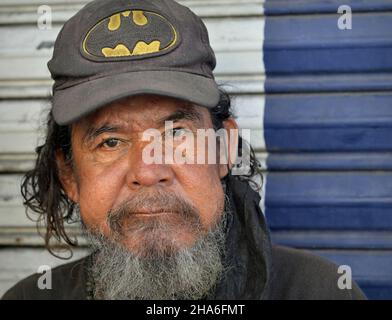 Älterer, robuster, obdachloser mexikanischer Mann mit grauem Bart und zerzaust behaartem Haar trägt eine abgenutzte Baseballmütze und blickt auf den Betrachter. Stockfoto