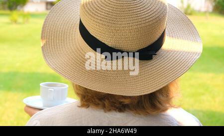 Frau in einem Strohhut hält eine Tasse heißes Getränk und trinkt Kaffee, sitzt am Morgen auf einer Sommerterrasse an einem sonnigen Tag. Das Konzept des Guten Stockfoto