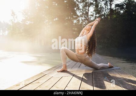 Frau, die an einem sonnigen Sommermorgen eine virabhadrasana-Übung, eine Kriegerpose und ein Training auf einer Holzbrücke in einem Park an einem Teich durchführt Stockfoto