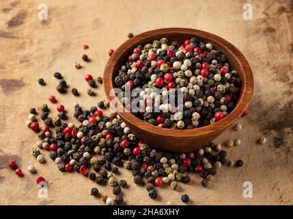 Pfeffer in einer Schüssel mischen. Schwarze, rote, weiße und grüne Pfefferkörner auf einem alten Schneidbrett. Stockfoto