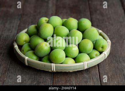 Unreife Pflaumen in einem Bambuskolander, der auf dem hölzernen Hintergrund platziert ist. Japanische Aprikosenfrüchte. Stockfoto