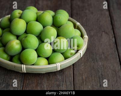 Unreife Pflaumen in einem Bambuskolander, der auf dem hölzernen Hintergrund platziert ist. Japanische Aprikosenfrüchte. Stockfoto