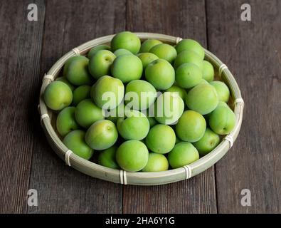 Unreife Pflaumen in einem Bambuskolander, der auf dem hölzernen Hintergrund platziert ist. Japanische Aprikosenfrüchte. Stockfoto