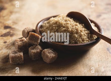 Brauner Zucker in einer Holzschüssel vor einem alten Holzhintergrund, daneben Zuckerwürfel. Stockfoto