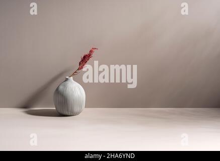 Minimalistische Stillleben-Szene. Kugelförmige Keramikvase und getrocknete Pflanzen. Beigefarbene Wand, Sonnenlicht und lange Schatten auf dem Hintergrund des Tisches. Trendy Stockfoto