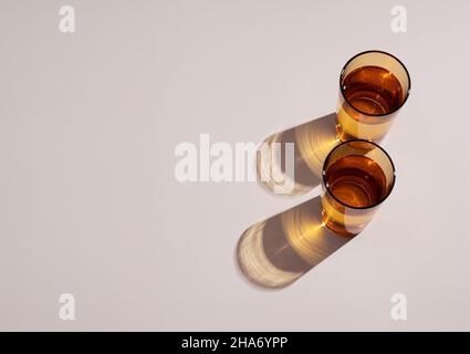 Dekorative Stillleben Szene im Sommer. Bernsteinglas mit Wasser, lange harte Schatten im Sonnenlicht. Beigefarbener Tischhintergrund. Flach liegend, Draufsicht. Stockfoto