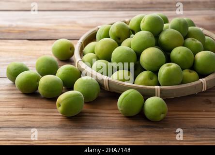 Unreife Pflaumen in einem Bambuskolander, der auf dem hölzernen Hintergrund platziert ist. Japanische Aprikosenfrüchte. Stockfoto