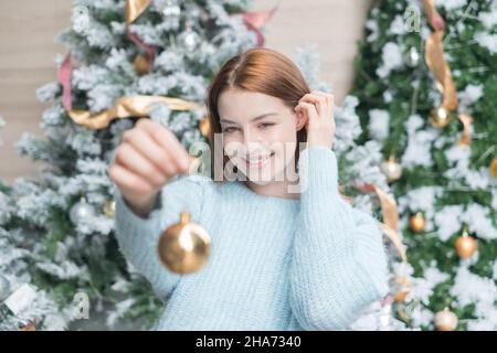 Neujahrs-Porträt der schönen kaukasischen jungen Frau in kuscheliger Wolle warm hellblau Pullover mit einem Goldball zur Dekoration weihnachtsbaum. Stockfoto