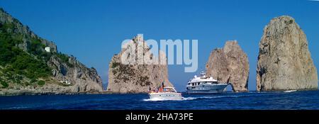 Boote, die zwischen der als Faraglioni de Mezzo bekannten Felsformation vor der Insel Capri, Italien, vorbeifahren, gelten als die ikonischste Sehenswürdigkeit der Insel Es ist eine Tradition, dass das Küssen eines Partners unter dem Bogen, während ein Boot durch sie fährt, dem betreffenden Paar Glück bringt. Das Schiff in der Mitte ist die Charter-Motoryacht IDOL, Stockfoto