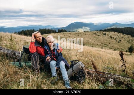 Ferientouristen, kaukasisches Paar Mann und Frau sitzen während eines Stopfens auf einem umgestürzten Baum. Wandern in den Bergen während des Urlaubs. Stockfoto