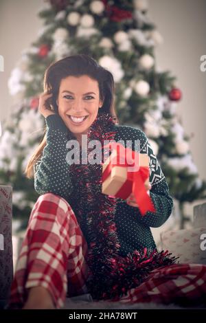 Ein junges schönes Mädchen genießt posiert für ein Foto, während halten ein weihnachtsgeschenk und sitzt auf dem Boden während der weihnachtsferien in einem angenehmen an Stockfoto