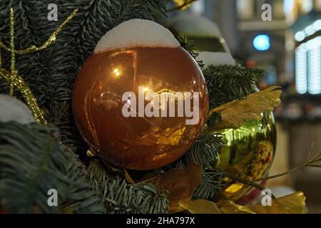 Auf der Straße befindet sich eine Tanne, die mit Neujahrsspielzeug geschmückt ist. Schnee bedeckt die Nadeln und festliche Kugeln. Die Kugeln spiegeln die Weihnachtsbeleuchtung wider Stockfoto
