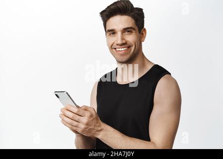 Lächelnder, gesunder Erwachsener mit männlichem Körper, der mit dem Smartphone steht, glücklich auf die Kamera schaut, mit der Handy-Anwendung, weißer Hintergrund Stockfoto