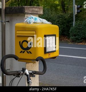 Auf einen gelben Briefkasten der Deutschen Post hat jemand einen Haufen Corona-Masken gelegt. Davor steht ein Fahrrad, der Lenker ist zu sehen. Stockfoto