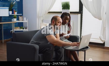 Junges interrassisches Paar, das mit einem Laptop und einem Notebook im Wohnzimmer ein Budget für Steuerzahlungen plant. Multiethnische Menschen berechnen Geld für Steuern. Mann und Frau überprüfen die Buchhaltung Stockfoto