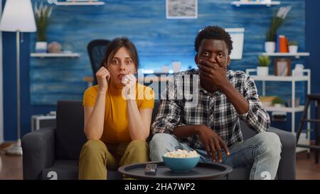 Interrassisches Paar, das sich im Wohnzimmer einen Theaterfilm im Fernsehen ansieht. Junge, multiethnische Partner sind schockiert, als sie auf der Couch auf die Kamera schauen. Ehemann und Ehefrau mit gemischter Rasse zu Hause Stockfoto
